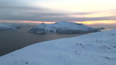 İnsansız hava aracının karlı bir dağın üzerinden uçup donmuş tepelerin ardında parlayan turuncu günbatımını gösteren bir tepe ve fiyort görüntüsü. Norveç 'te