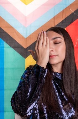 Gözleri kapalı genç bir kadının dikey portresi, yüzünün yarısını eliyle kapatıyor, trans gurur bayrağının önünde duruyor.. 