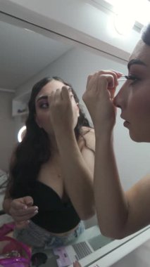 Aynadaki dikey yansıma, genç makyaj sanatçısının belden başa doğru dikkatli hareketlerle kırbaçladığını gösteriyor.. 