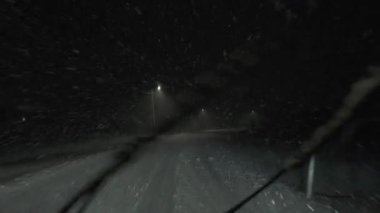 Uzaktaki sokak lambaları ve cam silecekleri kar temizlerken, bir arabanın içindeki yavaş çekim görüntüsü karlı bir yolda ilerliyor.. 
