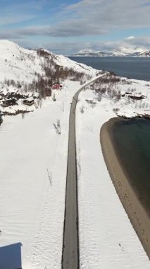 Karlı bir sahil ve yükselen dağ boyunca düz bir yolu takip eden dikey bir drone uçuşu ardında açık mavi gökyüzünün altında kırmızı bir arktik ev bırakıyor..