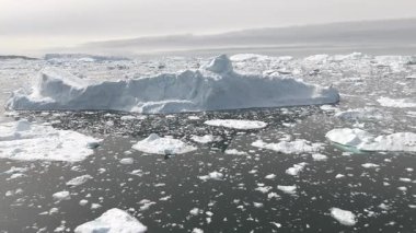 Yatay İHA panoraması bulutlu Arktik gökyüzünün altında sayısız buz parçasıyla çevrili devasa bir buzdağı etrafında döner..