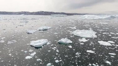 İnsansız hava aracı buzdağlarıyla dolu bir denizin üzerinde ilerliyor. Grönland 'daki kara parçası bulutlu yaz kutup gökyüzü altında kayboluyor..