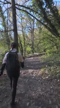 Gün ışığı aydınlık bir bahar gününü aydınlatırken, bir kadın açık hava kıyafetleri içinde orman yolunda yürüyor.