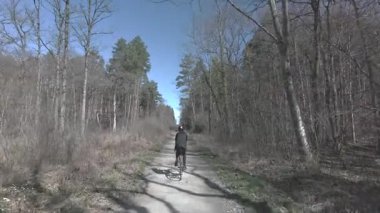 Güneş ışığı ilkbahar ağaçlarının arasından süzülürken bir kadın dar bir orman yolunda bisiklet sürüyor.