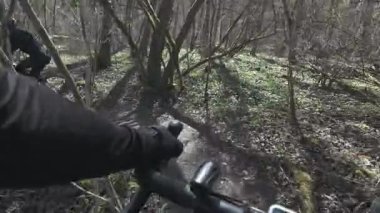 Nehir kenarındaki orman yolunda bisiklete binen ilk kişi görünür gidonu ve ileride başka bir binici daha var.