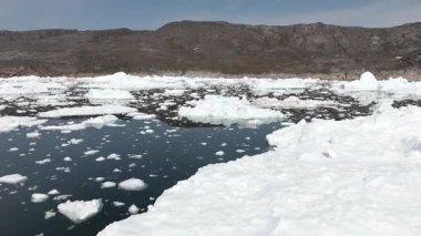 İleriye doğru hareket, Grönland kıtası ve bulutlu gökyüzünün arkaplanı çerçevelediği sakin denizde buz parçalarına yakın kalır.