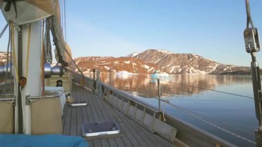 Tahta yelkenli sessizce karlı dağların yanından geçiyor Grönland 'ın sakin sularını yansıtıyor.