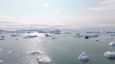İki kişi kayalık bir kıyı şeridinde durup kutup okyanusunda yüzen buzdağlarını seyrediyor.