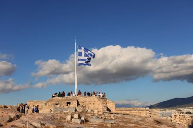 Atina, Yunanistan - 29 Eylül 2025: Atina 'daki Akropolis tepesinde bulutlu mavi bir gökyüzünün altında dalgalanan Yunan bayrağı, antik kent manzarasına hayran turistler.
