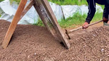 Havan ve duvar sıvası hazırlamak için ince parçacıkları çakıllardan ayırmak için ahşap bir ekranla kumu elleyen bir inşaat işçisine yaklaş..