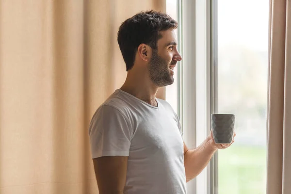 Skjuten av en ung man som tar en kopp kaffe medan han tittar ut genom fönstret hemma. — Stockfoto