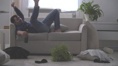 Kanepede oturan genç sakallı adam bira şişesini alır ve 4K yavaş çekim videosundaki şaşkınlıkla ona bakar. Akşamdan kalma olduğu için kendini kötü hissediyor..