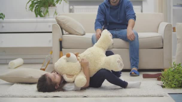 Samotne dziecko bawiące się na podłodze z misiaczkiem podczas gdy jej pijany tatuś jest z butelką na kanapie w teledysku 4k. — Wideo stockowe