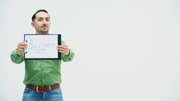 Funny young man standing holding the paper holder with word bisiness writen on it, showing it at the camera, pointing at it, playing, moving comically. — Stock Video