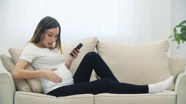 4k vídeo de mulher grávida sentada no sofá branco com telefone. — Vídeo de Stock