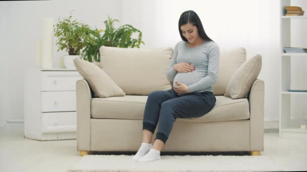 4k slowmotion video van zwangere vrouw zitten op witte bank en het aanraken van haar maag. — Stockvideo