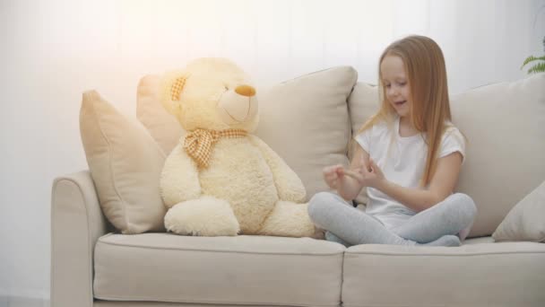 Vidéo ralenti 4k de petite fille en vêtements blancs jouant sur un canapé blanc avec ours en peluche. — Video