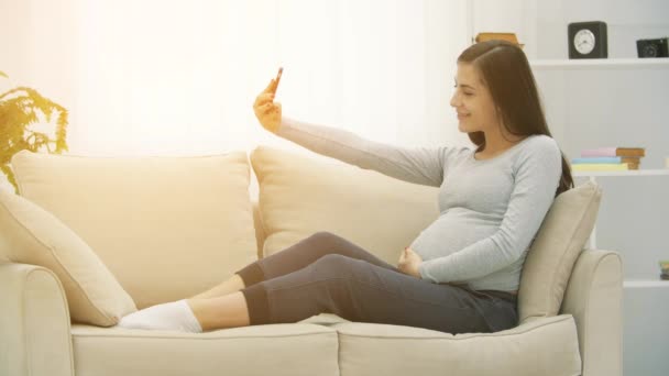 4k slowmotion video van zwangere vrouw zitten op een witte bank en het maken van selfie. — Stockvideo