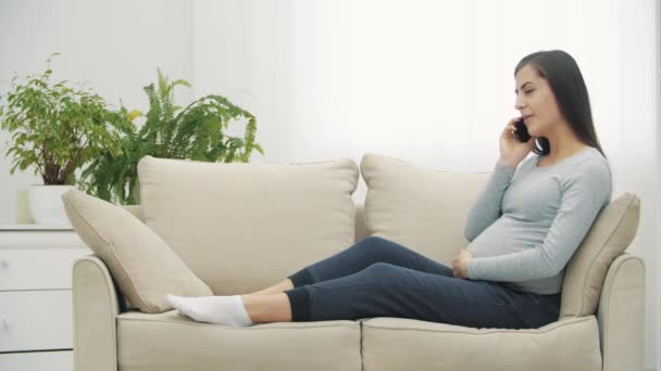 4k video of pregnant woman talking over the phone on white sofa. — ストック動画