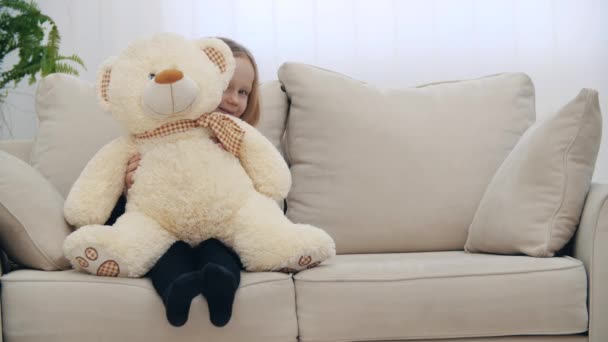 Mała uśmiechnięta dziewczynka siedzi na kanapie ze swoim pluszowym misiaczkiem w 4k wideo. — Wideo stockowe