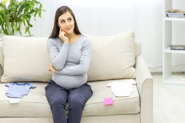 Foto von schwangerer Frau sitzt auf dem Sofa und kindische Kleidung in der Nähe. — Stockfoto