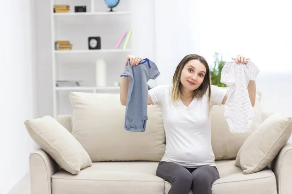 Foto de una mujer embarazada sentada en el sofá y mostrando ropa de niño. — Foto de Stock