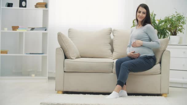 4k slow motion video de la mujer embarazada sentada en un sofá blanco y tocando su estómago. — Vídeos de Stock