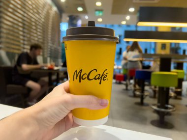 Poznan, Polonya - 11 Eylül 2025: Bir kişinin eli, hareketli bir fast food restoranı ortamında popüler markayı gözler önüne seren, tek kullanımlık parlak sarı bir McCafe kahve bardağını tutuyor.