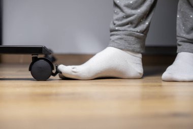 Beyaz bir çorabın içinde, siyah bir Caster tekerleğin fren mekanizmasını aktif olarak çalıştıran, pürüzsüz ahşap zeminde mobilya dengesini sağlayan, insan ayağının detaylı bir yakın çekimi.