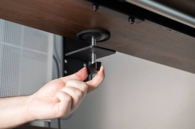 Siyah klemp düğmesini çeviren bir elin yakın plan görüntüsü, koyu ahşap bir masa başına masa aksesuarı takmak, çalışma alanı çözümleri için etkin ev ofis kurulumunu gösteren