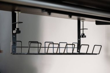 Koyu ahşap bir masanın altına yerleştirilmiş, temiz, dağınıklıktan arınmış ve üretken bir ofis ortamı sağlayan, parlak, siyah bir kablo düzenleyici sepetinin altında.