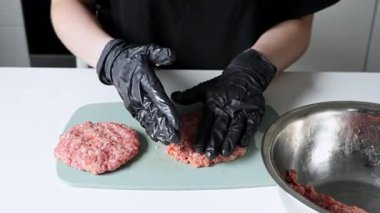 Açık yeşil bir kesme tahtasının üzerinde çiğ et köfteleri oluşturan siyah eldivenli kişilerin yakın plan çekimleri, temiz mutfak uygulamaları ve yemek hazırlığını vurguluyor. Büyüt