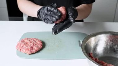 Siyah koruyucu eldivenler giyen ellerin yakın plan görüntüsü mükemmel şekillendirilmiş çiğ sığır burger köftesi yaratarak modern bir mutfakta güvenli ve dikkatli yemek kullanımını gösteriyor. Büyüt