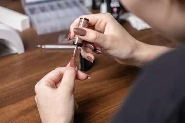 Kadının eli, tırnaklarına doğal kahverengi jel ojesi sürüyor. Güzellik tedavisini ve kişisel bakım ritüelini tasvir ediyor.