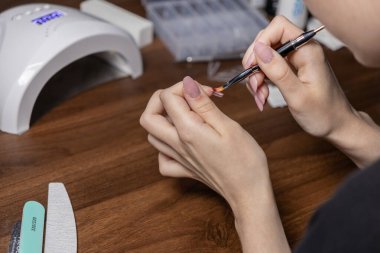 Çivi ucuna titizlikle poligel oymak için ince bir fırça kullanan ellerin detaylı görüntüsü, kişisel bir salon ortamında zarif ve uzun süreli bir manikür hazırlıyor.