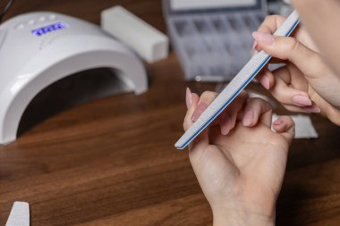 Açık pembe akrilik tırnakları şekillendirmek için bir tırnak törpüsü kullanan birinin ellerinin yakın çekimi, kişinin kişisel güzellik ayinini ve evde tırnak bakımını gösteriyor.