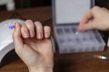 Bir kadının cilalanmamış elinin ve tırnaklarının yakın çekimi, kendi başına tırnak uzatmak için hazırlanması, iyileştirme lambasının yanındaki organizatörden üst form seçimini göstermesi.