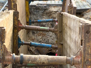 Mavi destekli paslı metal destekli çerçeveler, kazılmış bir siperin derin duvarlarını güçlendirerek, temel yeraltı tesisatı kurulumu veya bakım sırasında işçilerin güvenliğini sağlar.