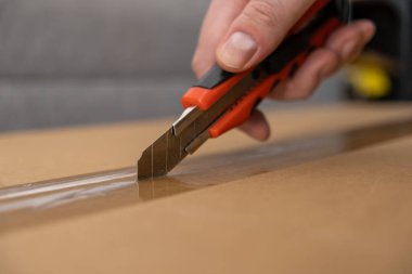 Kahverengi bir karton kutunun üzerinde açık bir bant dilimleyen kırmızı bir bıçakla bir insan eli, yeni teslimatları veya sevkiyatları açmanın pratik sürecini gözler önüne seriyor.