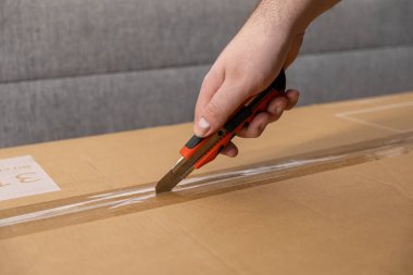 Bir insan elinin yakın plan görüntüsü büyük kahverengi bir karton kutunun üzerini tamamen temizler kırmızı bir bıçakla, paketin güvenli bir şekilde gelişini ve kutunun açılmasını temsil eder.