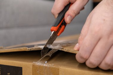 İnsan elinin bantlanmış kahverengi bir karton paketi açmak için bıçak kullanışının yakın plan görüntüsü e-ticaret emri alma sürecini gösteriyor.