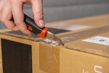 Büyük kahverengi bir teslimat kutusunun mührünü kırmak için keskin bir bıçakla e-ticareti ve mal alımını temsil eden bir kişinin elinin yakın çekimi.