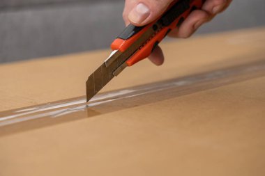 Bir insanın elinin yakın plan çekimi bir nakliye kutusundaki açık yapışkan bandı keserek eve teslimatı ve paketleri açmayı temsil ediyor.