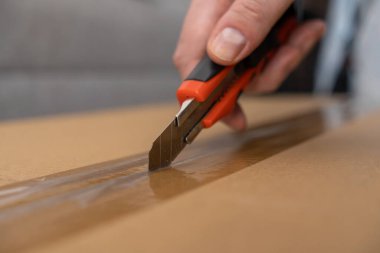 Parlak kırmızı bir alet bıçağını elinde tutan bir insan elinin yakın çekimi. Kahverengi bir karton kutunun şeffaf bandını sorunsuzca kesiyor. Hızlı teslimat ve paket erişimi için.