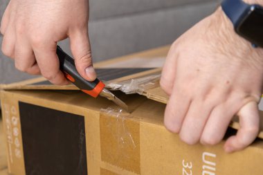 İnsan eliyle dikkatlice çekilmiş kahverengi bir karton paketi bıçak ile açarak ürün teslimatını ve yeni ürün elde etme başarısını temsil ediyor.