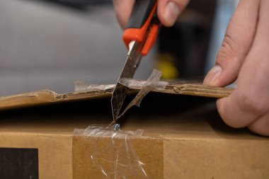 İnsan elinin yakın çekimi, sağlam kahverengi bir nakliye kutusunun güçlü ambalaj bandını keserek tüketici lojistiğini ve ürün varışını gösteriyor.