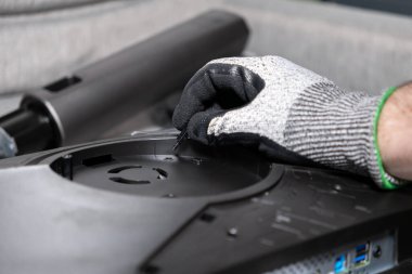 Güvenlik eldiveni içinde bir işçinin elinin yakın plan detayı bir LCD monitörünün arkasına küçük bir vida takarak, elektronik kurulumunda hassasiyet ve beceriyi gösterir