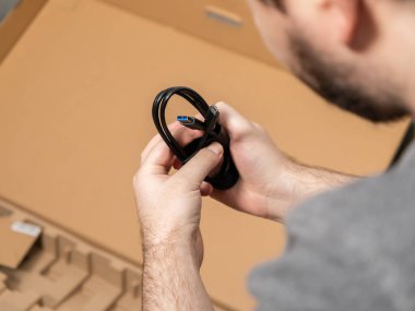 Karton bir kutudan sarmalanmış siyah bir USB veri transfer kablosu tutan bir adamın ellerinin yakın çekimi, yeni bilgisayar donanımını bağlama ve yapılandırma sürecini vurguluyor