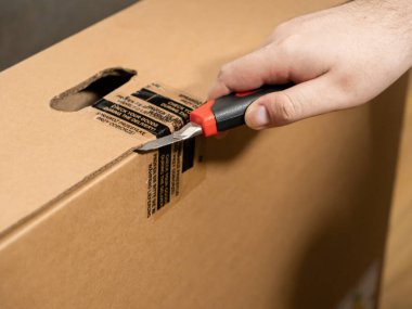 Mühürlü kahverengi bir karton paketi açmak için keskin bir bıçak kullanan bir kişinin elinin yakın plan çekimi, teslimat sırasında malları kontrol etme ihtiyacını vurguluyor